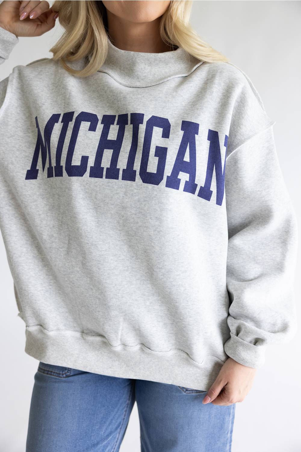 A woman wearing a light heather gray sweatshirt with 'MICHIGAN' printed across the chest in dark navy blue collegiate lettering, paired with blue jeans.