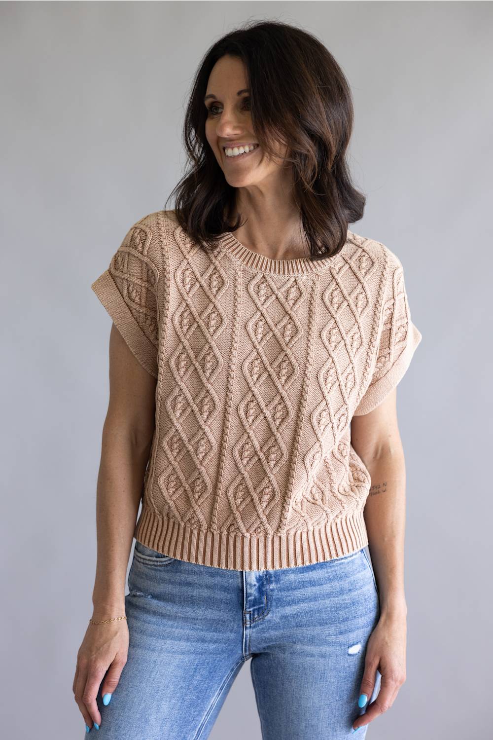A woman wearing a beige short-sleeve cable-knit sweater and blue jeans, standing against a plain light gray studio background.