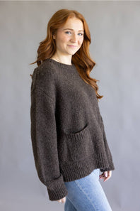 A woman with long red hair wearing a dark brown fuzzy knit sweater with a front pocket and light blue jeans, standing against a plain gray studio background.