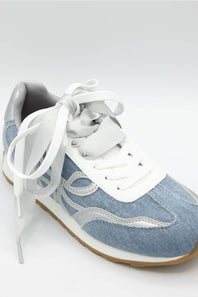 A light blue denim sneaker with white laces, white side panels, and a gum brown sole, photographed on a light gray background.