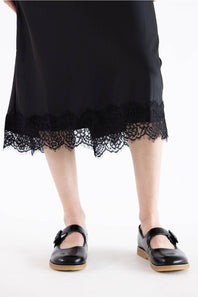 A person wearing a black midi skirt with a scalloped lace hem and black Mary Jane shoes with tan soles, shown from the waist down on a white studio background.
