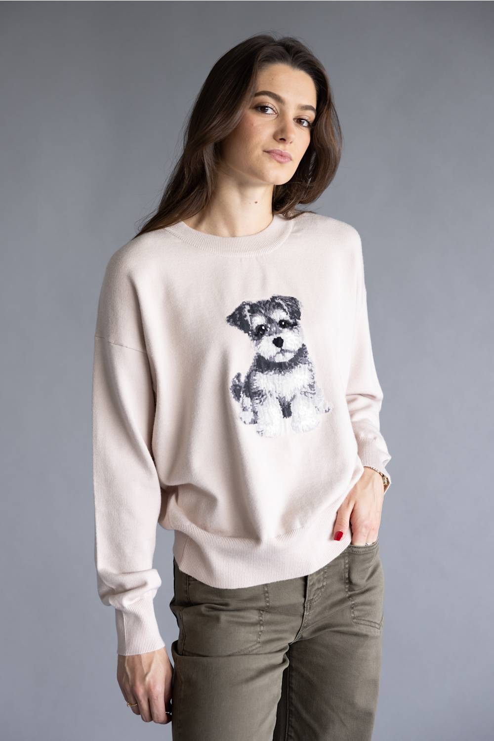 A woman wearing a light beige crewneck sweatshirt with a black and white graphic of a small dog on the front, paired with olive green pants, against a solid gray studio background.