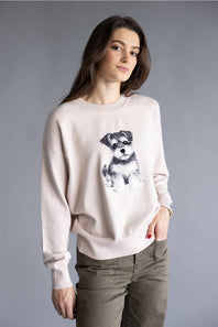A woman wearing a light beige crewneck sweatshirt with a black and white graphic of a small dog on the front, paired with olive green pants, against a solid gray studio background.