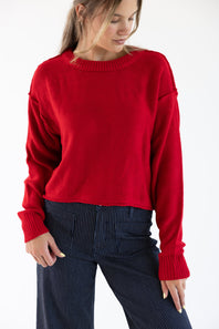 A woman wearing a red sweater with exposed seams styled with dark blue striped denim jeans standing in front of a white studio background