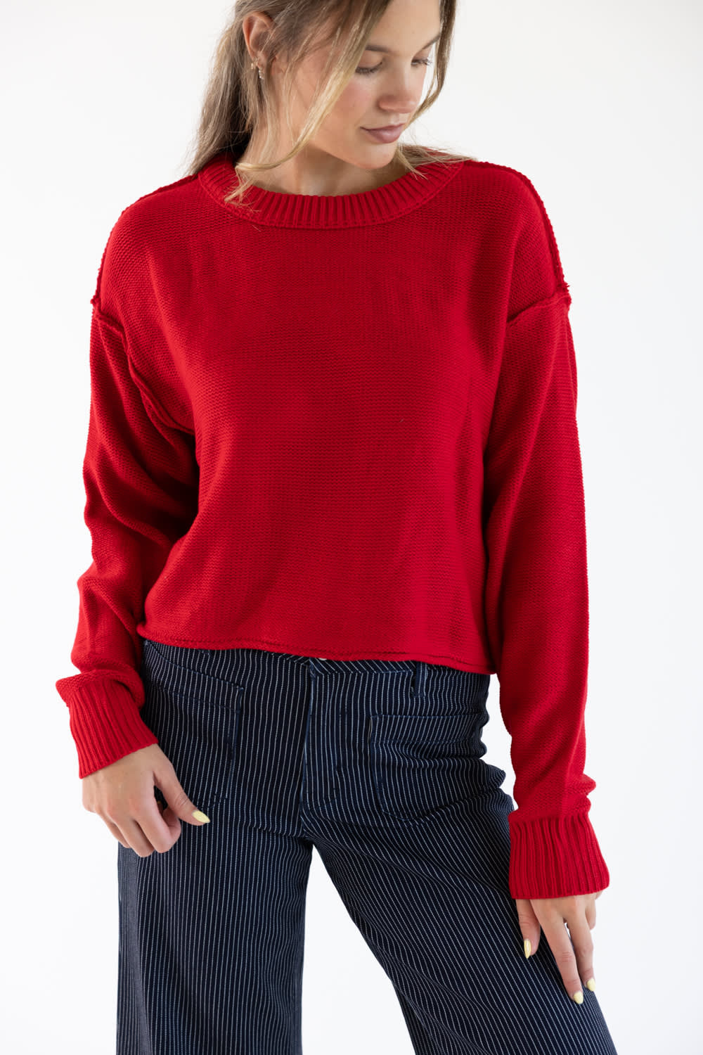 A woman wearing a red sweater with exposed seams styled with dark blue striped denim jeans standing in front of a white studio background