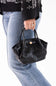 A person wearing a black patterned sweater and light blue jeans holds a small black crocodile-embossed handbag with double top handles and a gold-tone front accent against a plain white background.