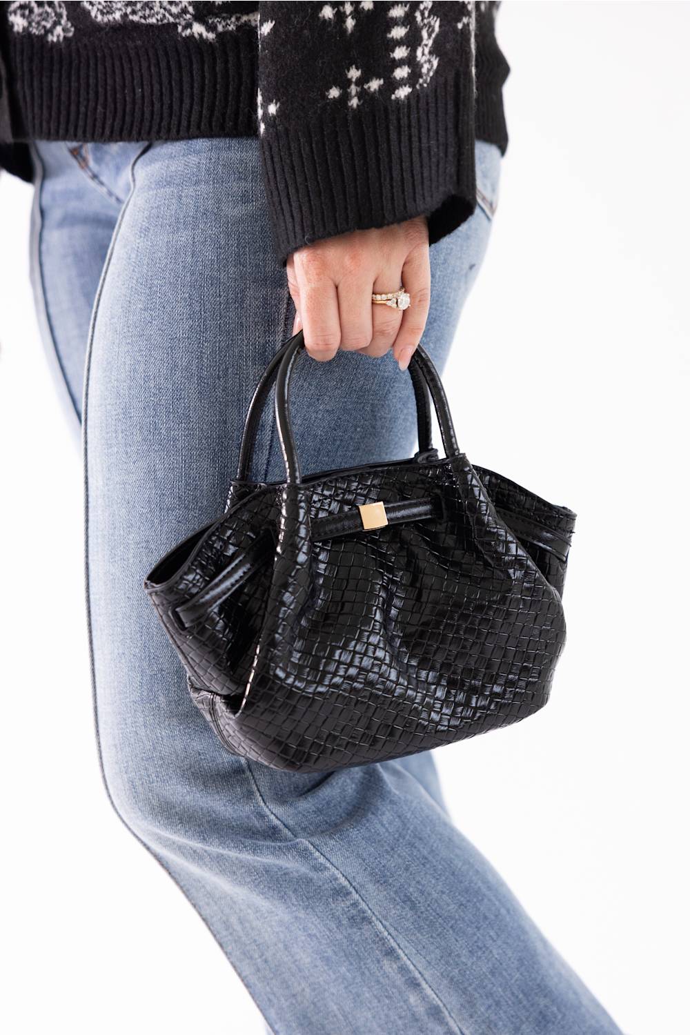 A person wearing a black patterned sweater and light blue jeans holds a small black crocodile-embossed handbag with double top handles and a gold-tone front accent against a plain white background.
