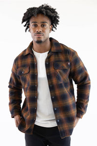 A man wearing a brown and navy plaid overshirt over a white t-shirt and black pants, photographed against a solid white background.