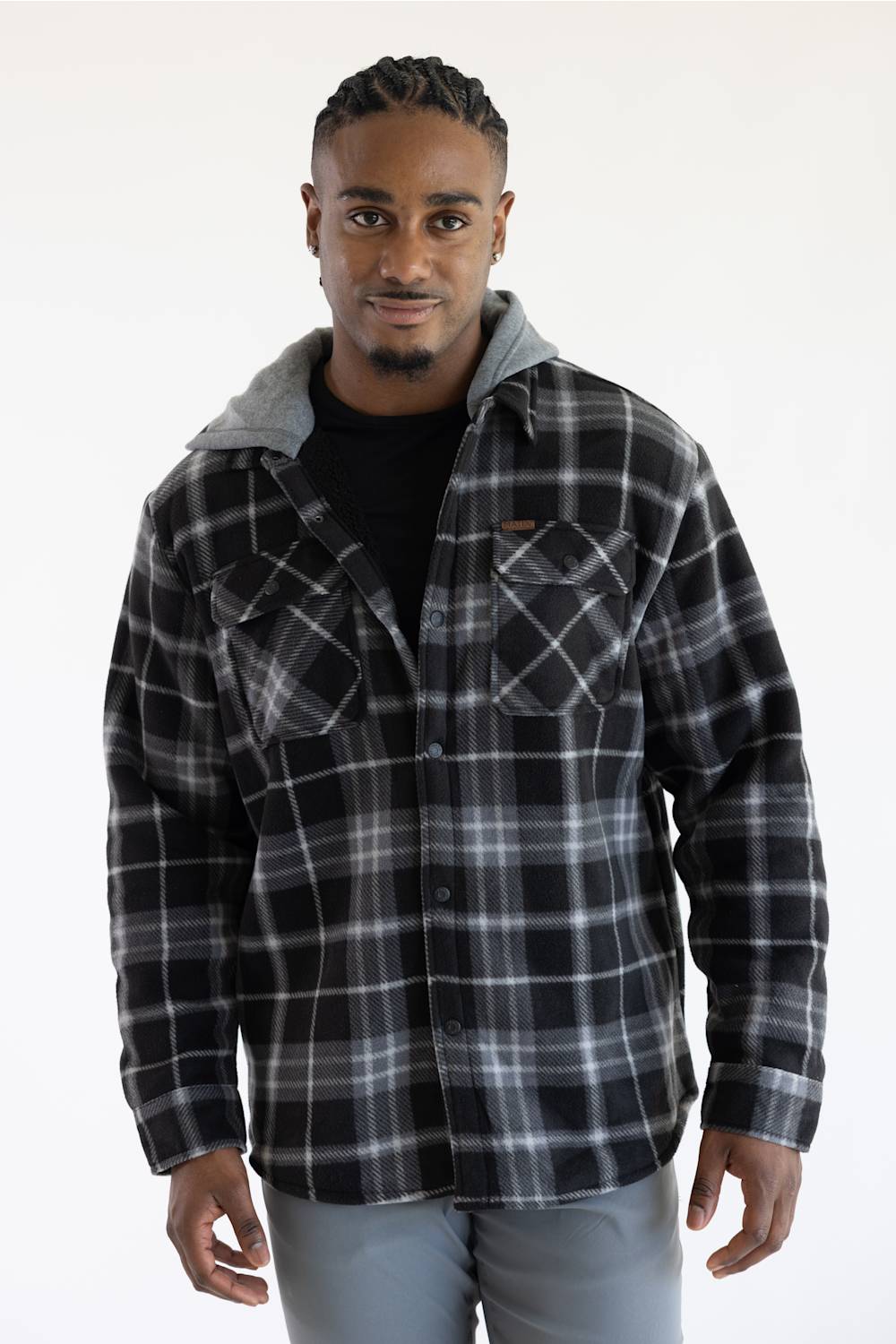 A man wearing a black and gray plaid hooded flannel shirt over a black t-shirt and gray pants, standing against a plain white studio background.