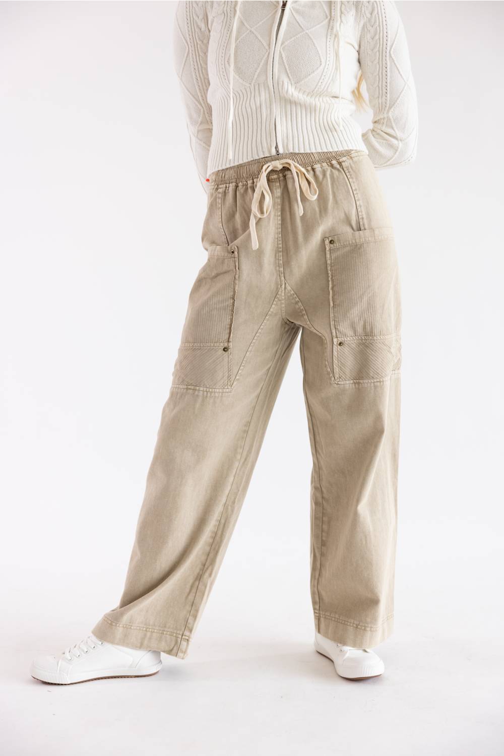 A person wearing beige wide-leg cargo pants with a drawstring waist, paired with a cream cable-knit sweater and white sneakers, shown from the chest down against a plain white studio background.