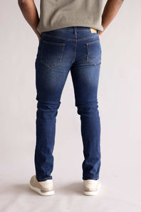 Person wearing blue jeans and a light-colored shirt on a plain background, showing two back pockets. The blue jeans are named Weatherproof Vintage Slim Jeans for Men in Silt Blue.