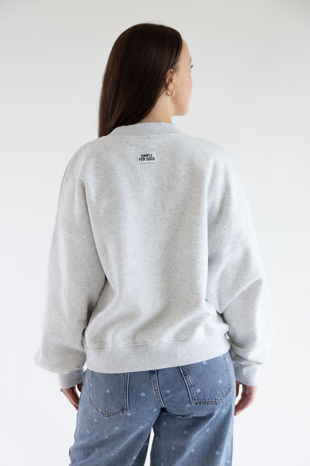 A woman shown from the back wearing a light gray heather crewneck sweatshirt and blue jeans with small polka dot details, against a plain white studio background.