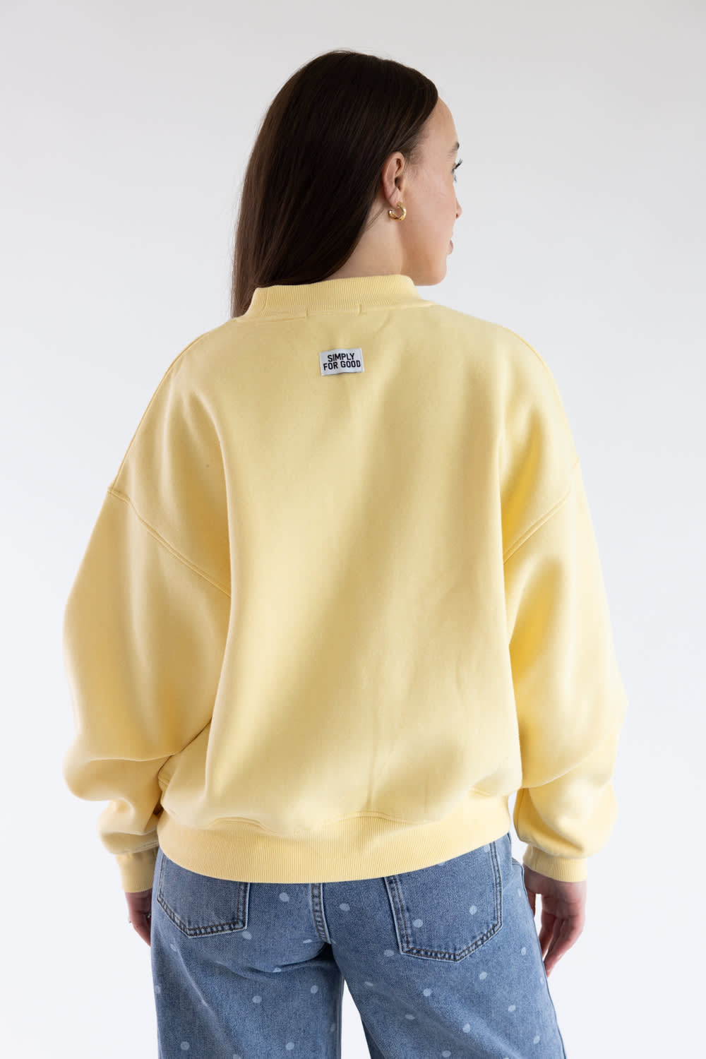 A woman viewed from the back wearing a pale yellow crewneck sweatshirt and blue jeans with small white star details, set against a plain light gray studio background.