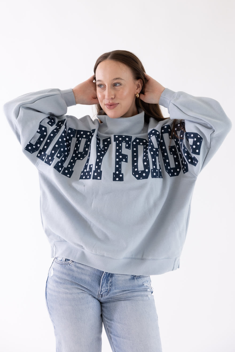 A woman modeling a light blue sweatshirt with 'SIMPLY FOR GOOD' written across the back in dark navy letters with white polka dots, paired with light blue jeans and white sneakers, against a plain white background.
