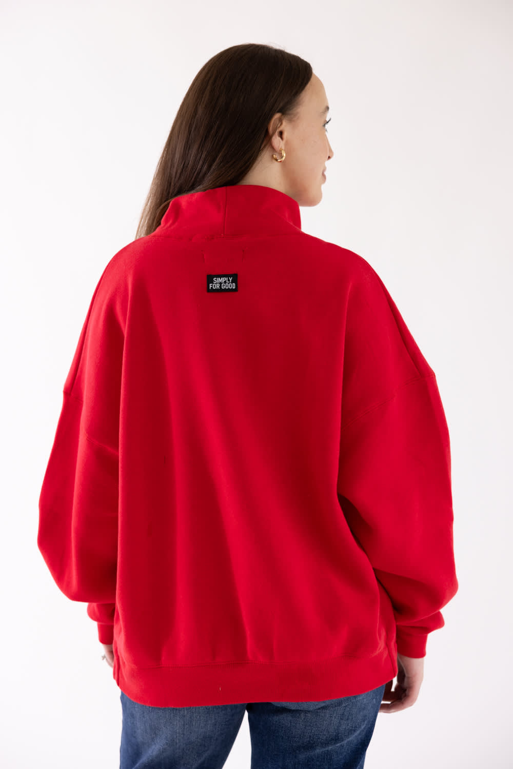 A woman shown from the back wearing a bright red sweatshirt with a small black brand patch on the upper back, paired with blue jeans against a plain white studio background.