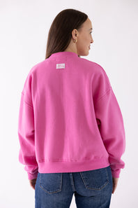 A woman shown from the back wearing a bright pink crewneck sweatshirt and blue jeans against a plain white studio background.