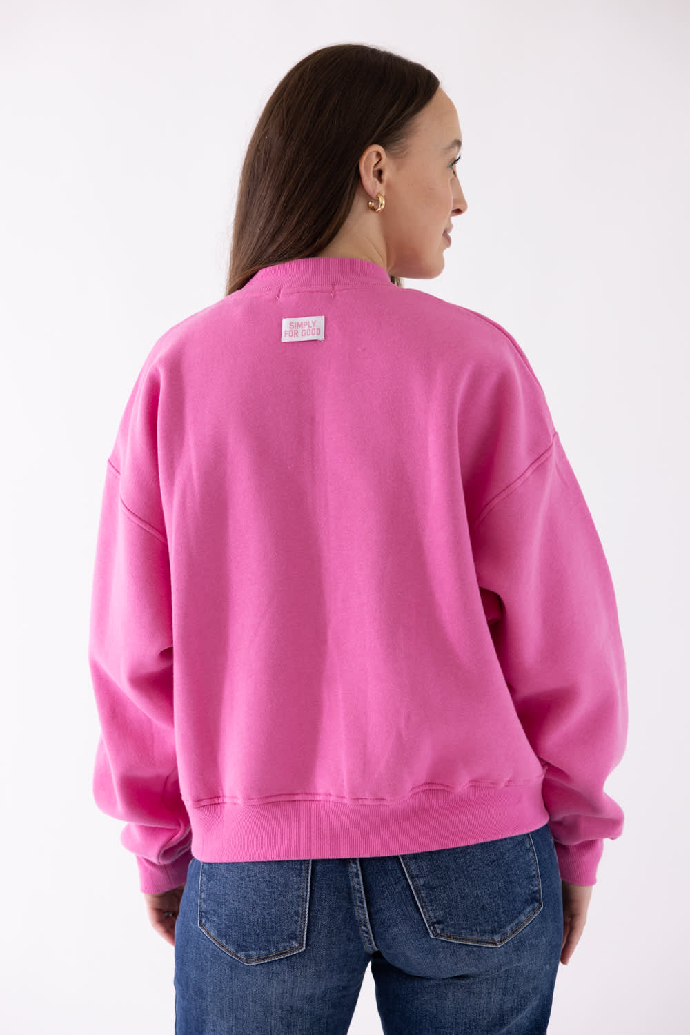 A woman shown from the back wearing a bright pink crewneck sweatshirt and blue jeans against a plain white studio background.