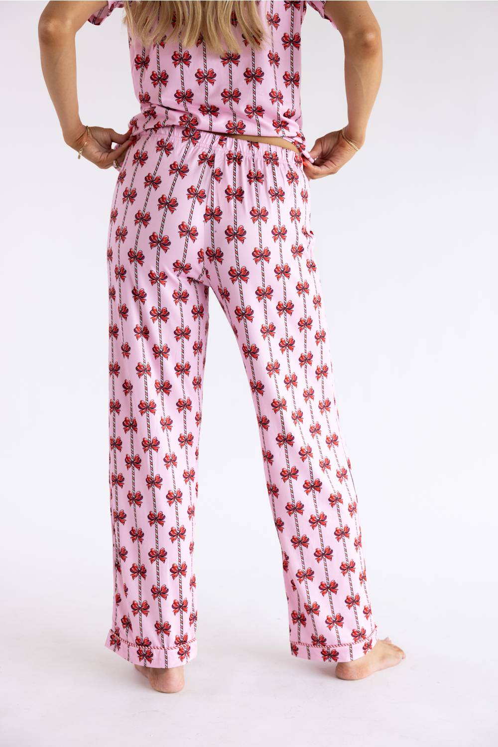 A woman wearing a pink two-piece pajama set with a red bow pattern, shown from the back against a plain white background.