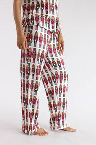A model wearing white wide-leg pajama pants covered in a colorful nutcracker soldier pattern, shown from the waist down against a plain white studio background.