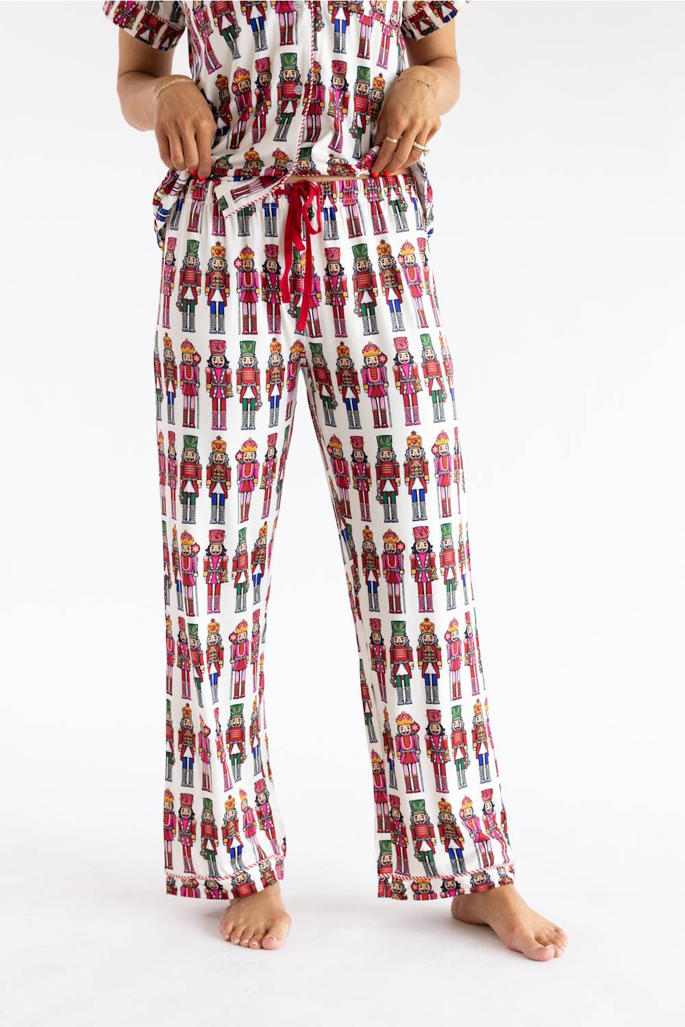 A person wearing a matching pajama set with a white base and an all-over colorful nutcracker soldier print, including a short-sleeve top and drawstring pants, photographed against a plain white studio background.