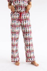 A person wearing a matching pajama set with a white base and an all-over colorful nutcracker soldier print, including a short-sleeve top and drawstring pants, photographed against a plain white studio background.