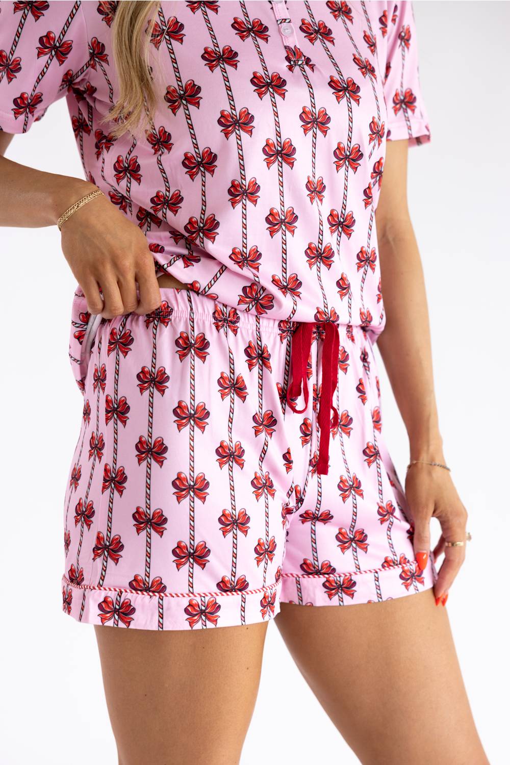 A woman wearing a pink short-sleeve romper with a red and black bow print, featuring a drawstring waist and matching shorts, shown against a plain white background.