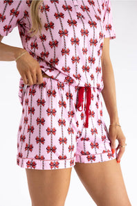 A woman wearing a pink short-sleeve romper with a red and black bow print, featuring a drawstring waist and matching shorts, shown against a plain white background.