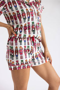 A model wearing a matching short-sleeve button-up shirt and shorts set in white with a colorful nutcracker soldier pattern, photographed against a plain white background.