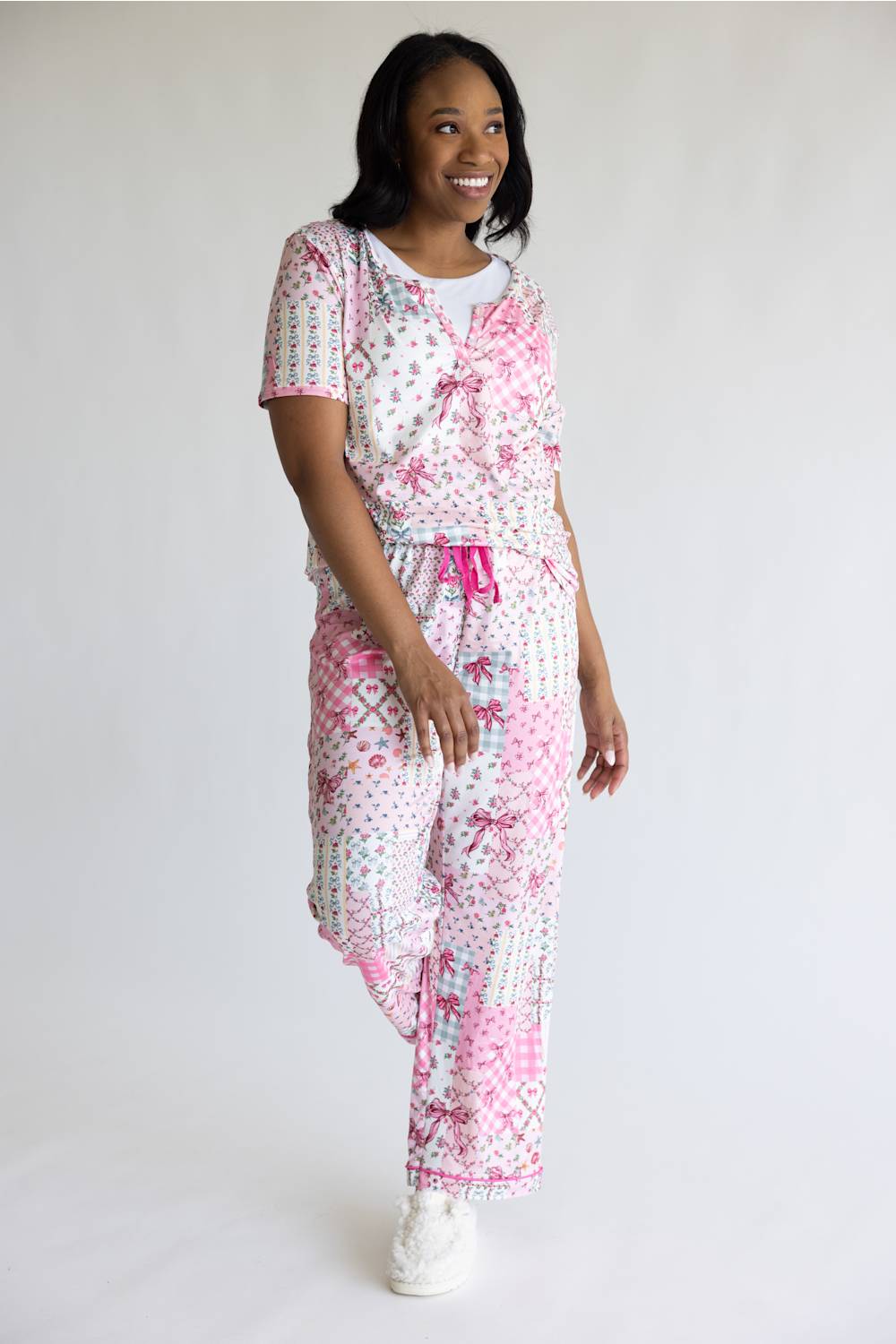 A woman modeling a two-piece pajama set featuring a pink and white floral print, consisting of a short-sleeve top with a tie detail and matching pants, against a plain white studio background.