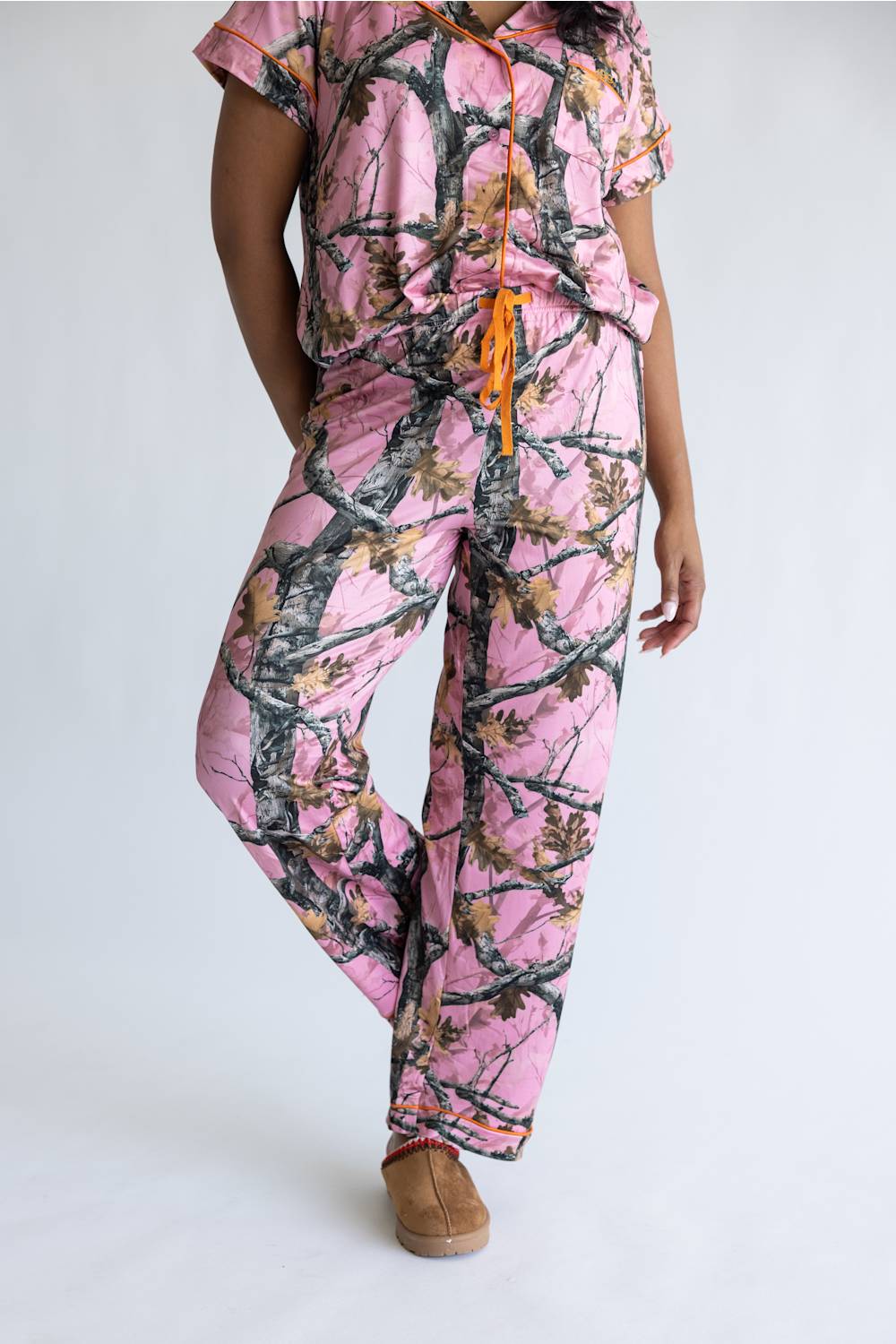 A person wearing a matching short-sleeve top and pants pajama set in a pink camouflage print with brown tree branches and an orange drawstring, paired with tan slip-on shoes, against a plain white studio background.