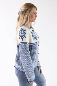 A woman shown in side profile wearing a blue and cream knit cardigan with snowflake patterns on the chest, paired with light blue jeans, against a plain white studio background.