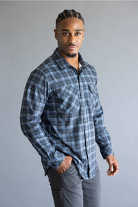 A man wearing a long-sleeve button-down plaid shirt in shades of blue and gray, paired with gray pants, standing against a solid gray studio background.
