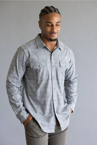 A man wearing a light gray and white plaid long-sleeve button-down shirt with gray pants, standing against a solid gray studio background.