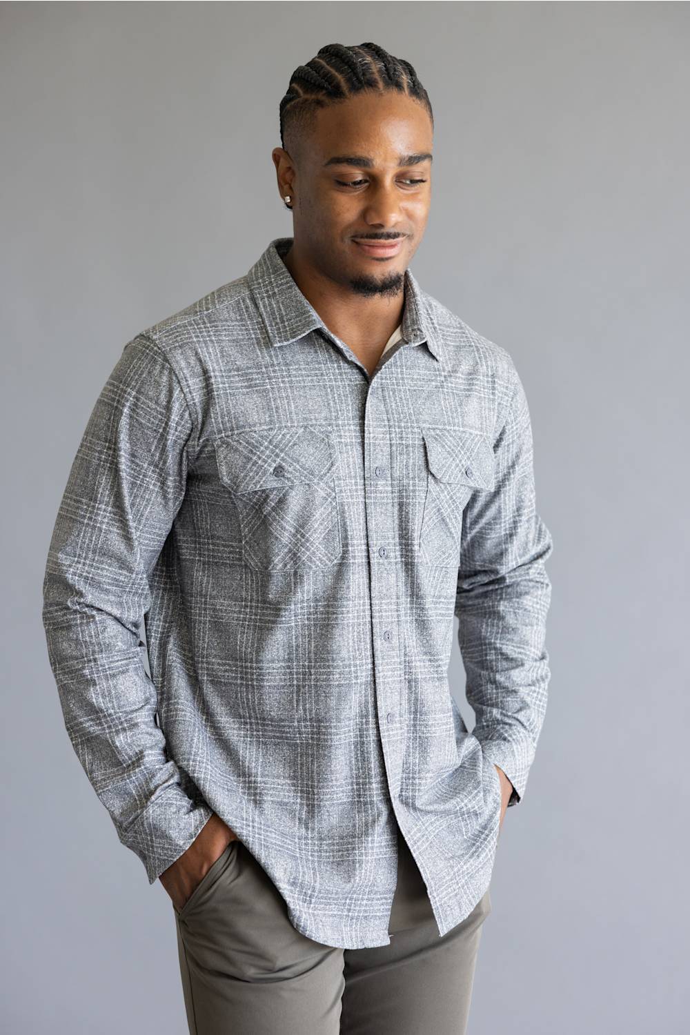 A man wearing a light gray and white plaid long-sleeve button-down shirt with gray pants, standing against a solid gray studio background.