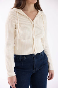 A woman wearing a cream-colored ribbed knit zip-up hoodie with a hood and drawstrings, paired with dark blue jeans, against a plain white background.