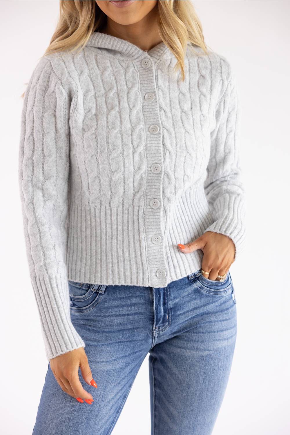 A woman wearing a light gray cable-knit button-front cardigan sweater with a hood, paired with blue jeans, shown against a plain white background.