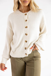 A woman wearing a cream-colored knit cardigan sweater with brown wooden buttons, paired with olive green pants, against a plain white background.