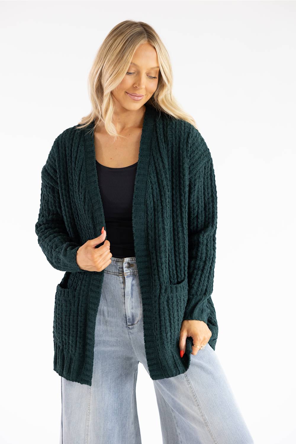 A woman wearing a dark forest green chunky cable-knit cardigan with front pockets, styled over a black top and light-wash blue jeans, against a plain white background.