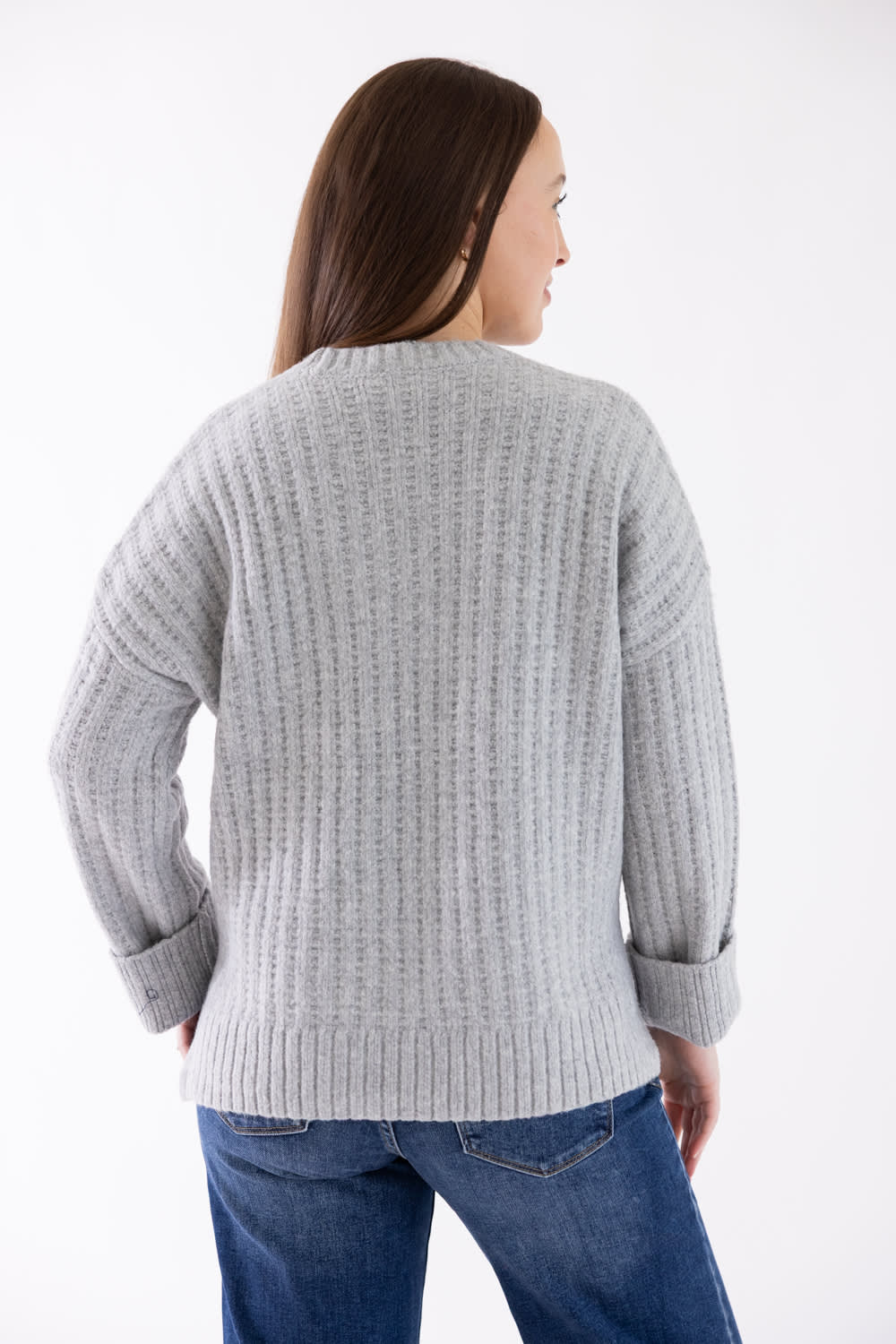 A woman shown from the back wearing a light gray ribbed knit sweater and blue jeans against a plain white studio background.
