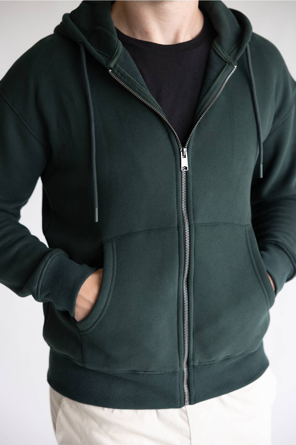 A man wearing a dark forest green zip-up hoodie with a black t-shirt underneath and off-white pants, shown from the neck down against a plain light background.