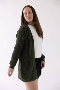A woman modeling a dark olive green waffle-knit open-front cardigan over a white top and black skirt, photographed against a plain white studio background.