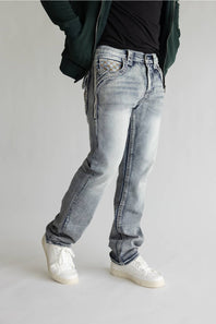 A person wearing light-wash blue jeans with decorative embroidered details on the back pockets, styled with a black t-shirt, dark green jacket, and white sneakers against a light gray studio background.