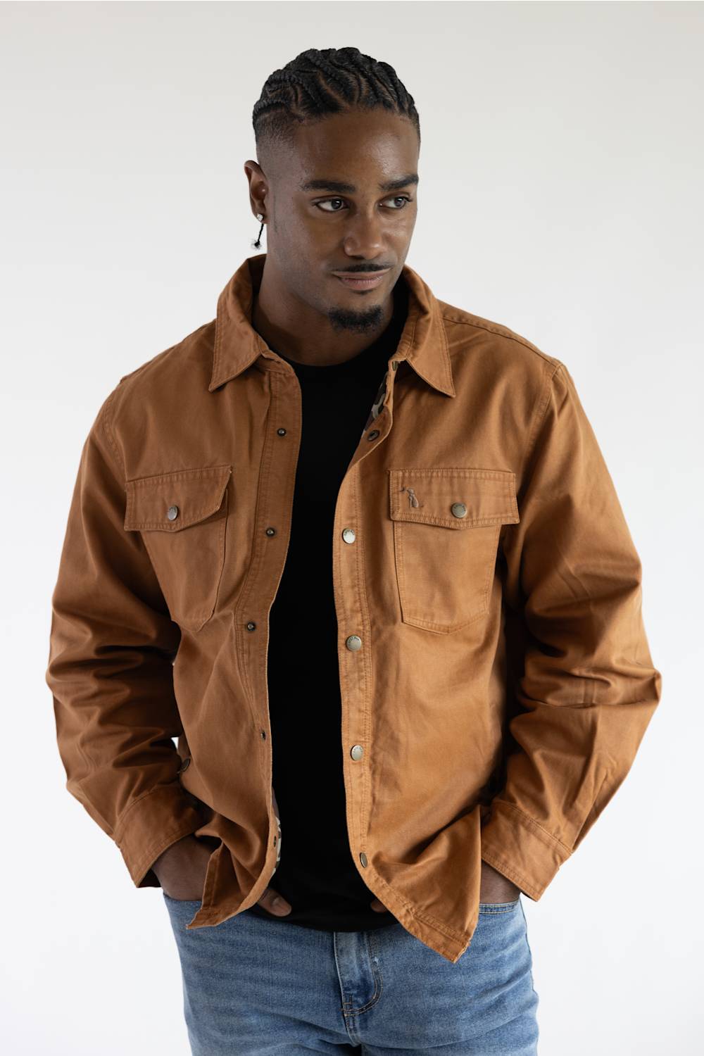 A man wearing a brown button-up overshirt jacket over a black t-shirt and blue jeans, photographed against a plain white studio background.