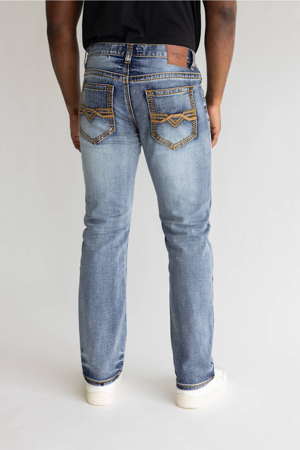 Rear view of a person wearing light-wash blue jeans with decorative brown contrast stitching on the back pockets, paired with a black top and white sneakers, against a light gray studio background.
