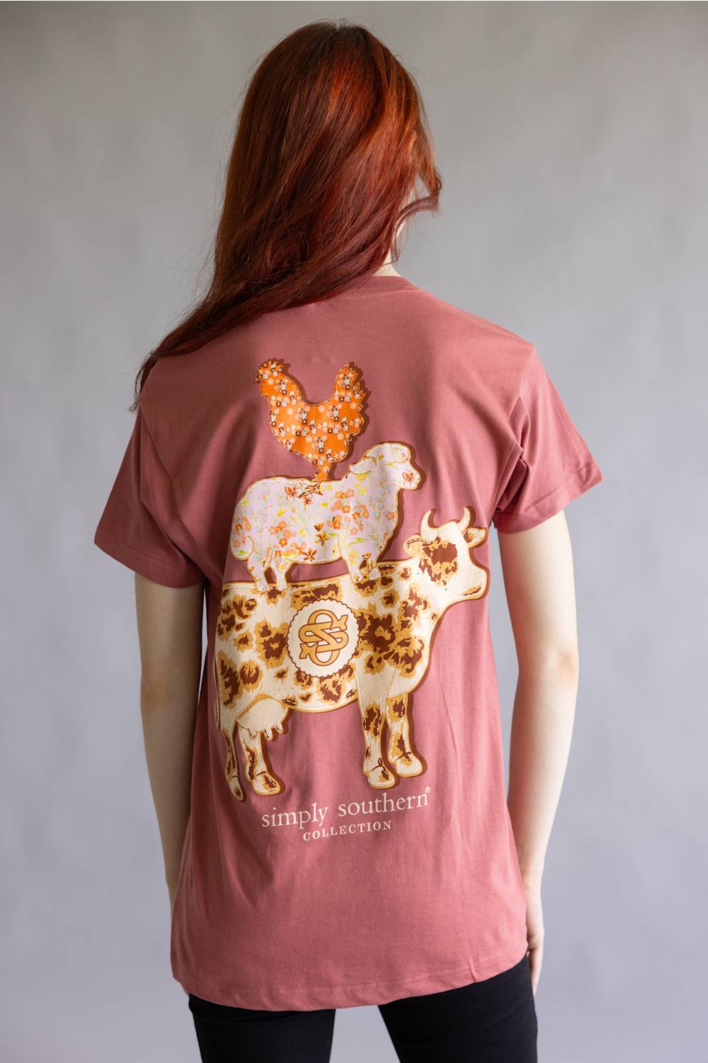 A woman viewed from behind wearing a dusty rose pink t-shirt with a large graphic of two decorated sheep and a butterfly on the back. The text 'simple southern collection' is printed below the graphic.