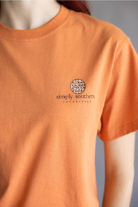 A close-up of a person wearing a short-sleeve crewneck t-shirt in a warm peach-orange color. The shirt features a small circular logo on the left chest with the text 'simply southern COLLECTION'.