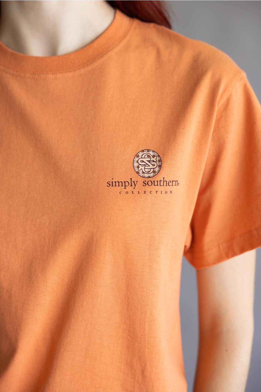 A close-up of a person wearing a short-sleeve crewneck t-shirt in a warm peach-orange color. The shirt features a small circular logo on the left chest with the text 'simply southern COLLECTION'.