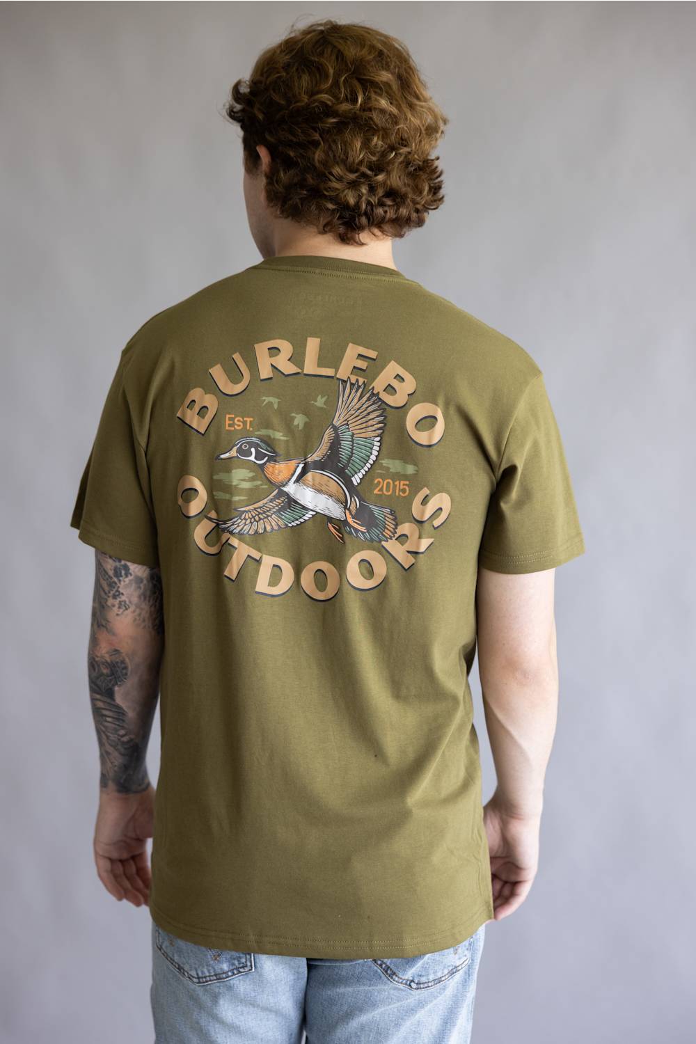 A man viewed from behind wearing an olive green t-shirt with a large graphic on the back featuring a duck and the text 'BURLEBO OUTDOORS'.