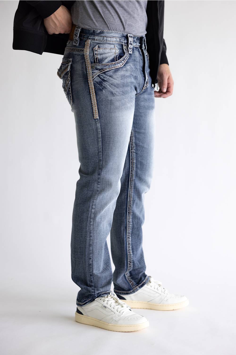 A person wearing light-wash blue jeans with decorative contrast stitching, a gray t-shirt, a black jacket, and white sneakers, shown from the waist down against a plain white studio background.