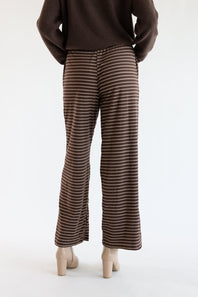 A person shown from the back wearing brown wide-leg pants with thin horizontal beige stripes, a dark brown sweater, and beige block-heel mules against a plain white studio background.